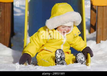 Toddler baby boy rides down a slide playing on a winter playground. A child in a yellow jumpsuit on a children's slide in the snow. Kid age one year e Stock Photo