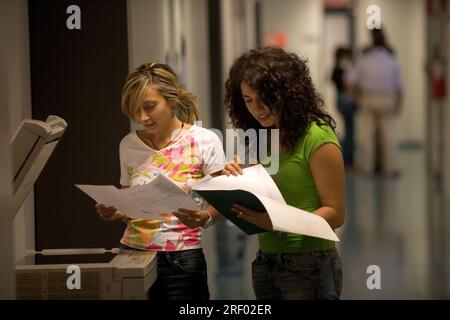 Two women reviewing documents beside a copier in a hallway. Stock Photo