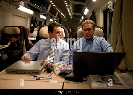 Two men in conversation on a train, using laptops and surrounded by passengers reading or resting. Stock Photo