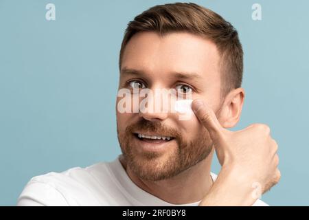 Portrait of bearded handsome man applying moisturizing cream on face skin looking at camera. Stock Photo