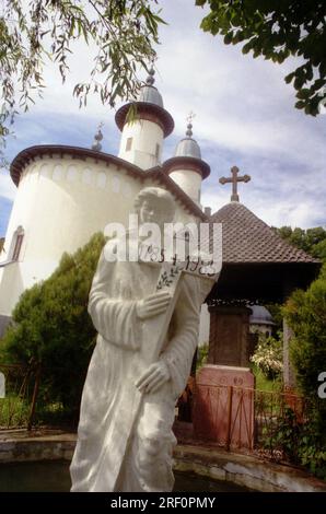 Varatec Monastery, Neamt County, Romania, 1999. A statue of an angel holding a cross on the grounds of the first wooden church of the monastery (built 1785). In the back, the 'Dormition of the Virgin Mary' church. Stock Photo