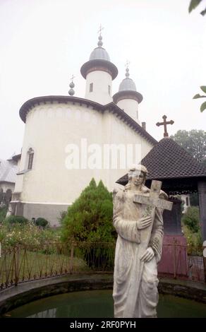 Varatec Monastery, Neamt County, Romania, 1999. A statue of an angel holding a cross on the grounds of the first wooden church of the monastery (built 1785). In the back, the 'Dormition of the Virgin Mary' church. Stock Photo