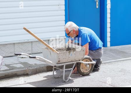 Elderly builder mason tile foundation of house. Working tiler at work outside on summer day. Contractor works at production facility. Stock Photo
