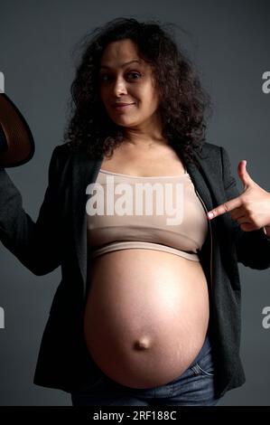 Multiethnic pregnant woman, gravid expectant mother pointing finger at her big pregnant belly in the ninth month of carefree happy pregnancy, smiling, Stock Photo