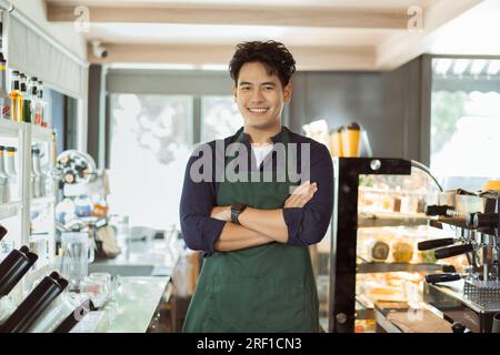 Young Asian male barista in apron having arms folded and smiling while looking at camera with copy space Stock Photo