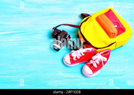 Pair of tiny red canvas shoes, colorful backpack and a toy camera arranged on a blue wooden background with copy space. Creative back-to-school or tra Stock Photo