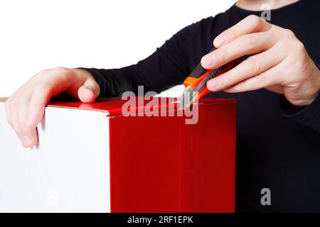Person wearing a black long-sleeve shirt skillfully cutting tape on a large red and white box using a utility knife. Packaging projects, office suppli Stock Photo