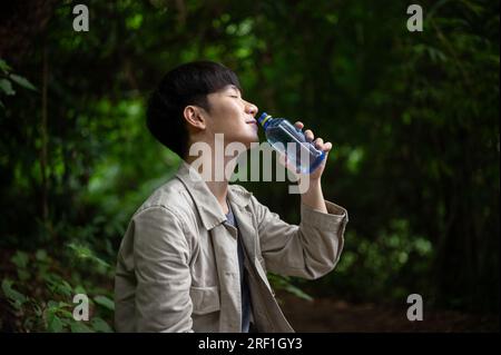 Rear view of a happy and relaxed Asian male hiker sits on a rock, eyes closed, drinking water from a bottle, while resting during hiking in the forest Stock Photo