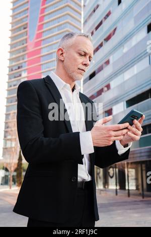 Vertical. Middle aged businessman using a smartphone to check his stock shares. Well dressed successful business person typing with a cellphone messaging app at workplace. Mature executive with phone. High quality photo Stock Photo