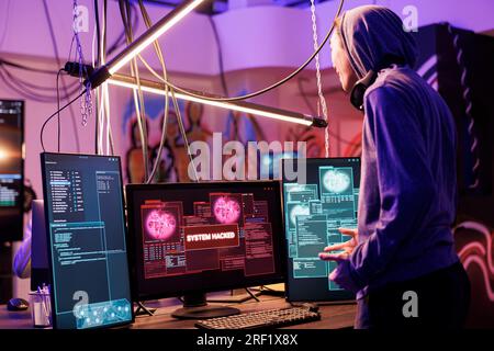 Happy hacker successfully breaking into website using malicious software. Cheerful smiling criminal in hood hacking computer network system in abandoned warehouse at night Stock Photo