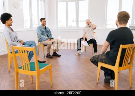 Remote portrait of professional male psychologist having conversation with multiethnic addicted people during therapy session, sitting in circle Stock Photo