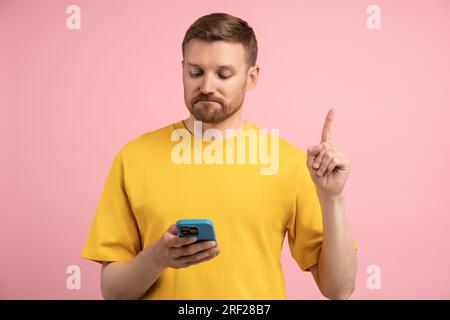 Sad upset man pointing index finger up looking in smartphone on pink background. Stock Photo