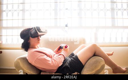 Gamer playing on vr headset with video game controller sitting on sofa Stock Photo