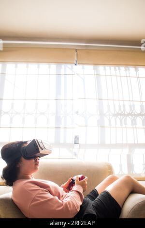 Gamer playing on vr headset with video game controller sitting on sofa Stock Photo