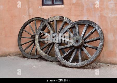 Three wooden wagon wheels leaning against orange beige brick wall. Stock Photo
