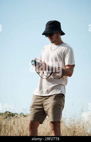 A portrait of a man in his twenties, casually dressed in a white t-shirt, beige shorts, sunglasses, and a black sailor cap, with a classic camera slun Stock Photo