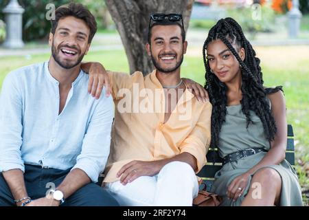 Two Caucasian men and a beautiful African-American woman with curly, braided hair sitting on a park bench, smiling as they pose for a photo. Stock Photo