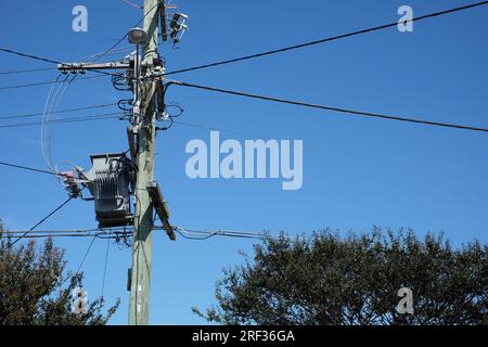 Power lines and a copper-treated, green wooden power pole with an elevated electricity transformer, street light, NBN cables against a clear blue sky Stock Photo