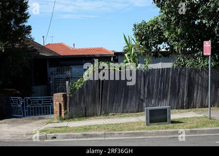 A TV sits on the edge of a grass footpath in the sun in front of a weathered wooden fence, a driveway with old iron gates in a western Sydney suburb Stock Photo
