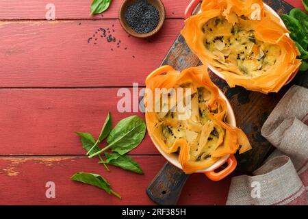 Filo pies with soft feta cheese and spinach in ceramic molds on old red wooden table backgrounds. Filo portions pies. Small Baked Spanakopita pies. To Stock Photo