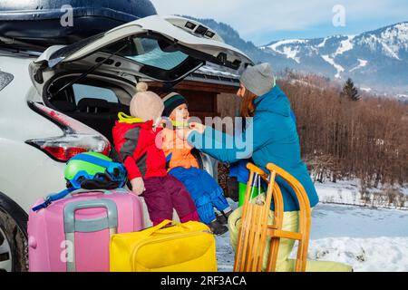 Mother help dress kids sitting in car trunk over snow mountain Stock Photo
