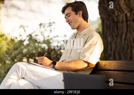 Millennial man student sitting on bench, doing homework Stock Photo