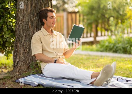 Carefree young guy sitting under tree at garden, reading book Stock Photo