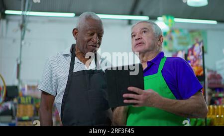 Two happy diverse senior staff of frocery store checking inventory operations with tablet device, older employees of supermarket laughing and smiling Stock Photo
