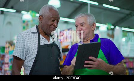 Two happy diverse senior staff of frocery store checking inventory operations with tablet device, older employees of supermarket laughing and smiling Stock Photo