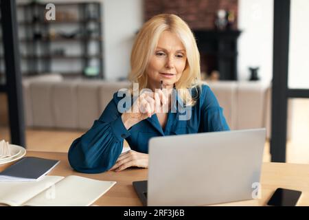 Distance work. Mature female freelancer working on laptop, sitting at desk in home office looking at screen and thinking Stock Photo