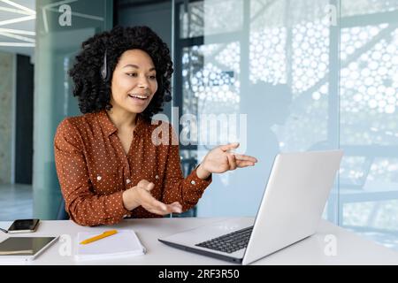 Young beautiful woman talking to customers remotely, african american female worker smiling using headset phone and laptop for online consultation, customer service. Stock Photo