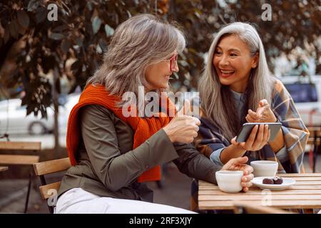 Cheerful senior Asian lady shows phone to grey haired friend at table with coffee and candies Stock Photo