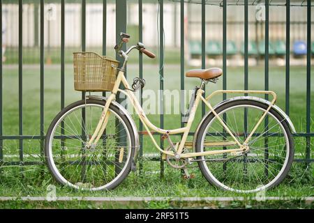 Vintage bicycle standing on the grass near the fence. Yellow retro bicycle with wicker basket. Stock Photo