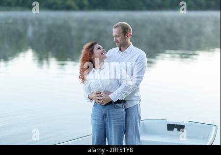 Romantic Caucasian man hugs Latin woman, couple enjoys time standing in boat, sailing in water, look at each other. Happy smiling lovers wear casual Stock Photo