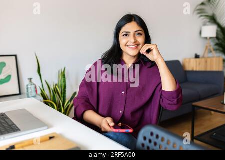 Young adult indian woman holding mobile phone while working with laptop at home Stock Photo