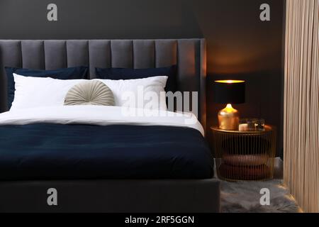Comfortable bed with cushions, lamp and different decor on bedside table in room. Stylish interior Stock Photo