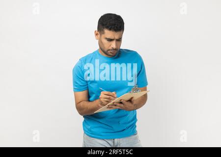Portrait of serious concentrated unshaven man wearing blue T- shirt standing with clipboard, writing something on paper, making to do list. Indoor studio shot isolated on gray background. Stock Photo