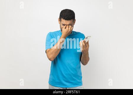 Portrait of tired sad unshaven man wearing blue T- shirt standing holding smart phone in hands, feels pain in his eyes, looking at screen long hours. Indoor studio shot isolated on gray background. Stock Photo