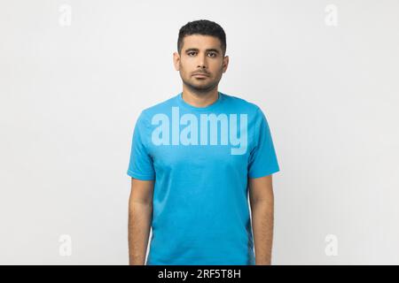 Portrait of serious strict handsome unshaven man wearing blue T- shirt standing looking at camera, having concentrated expression. Indoor studio shot isolated on gray background. Stock Photo