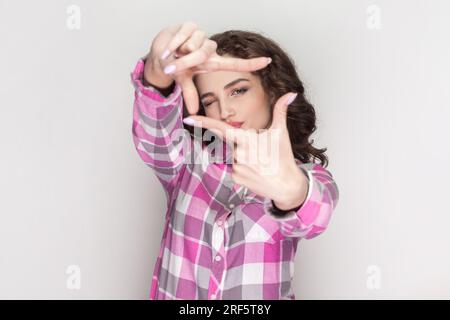 Happy ambitious woman with curly hair aims only success, makes hand frames searches perfect angle smiles broadly, gazes at camera through hands. Indoor studio shot isolated on gray background. Stock Photo