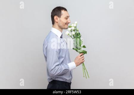 Side view portrait of smiling happy attractive man standing with bouquet and smelling flowers, enjoying pleasant aromat, wearing light blue shirt. Indoor studio shot isolated on gray background. Stock Photo