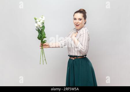 Side view portrait of excited woman wearing striped shirt and green skirt holding bouquet of white flowers, having pleasant present. Indoor studio shot isolated on gray background. Stock Photo