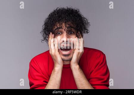 Portrait of shocked amazed bearded man with curly hair, looking in mirror, shocked from his hairstyle, keeps palms on cheeks, wearing red jumper. Indoor studio shot isolated on gray background. Stock Photo