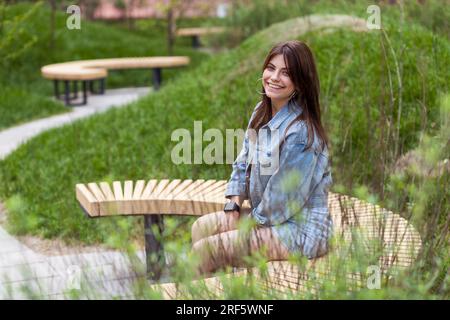 Side view portrait of extremely happy woman wearing denim jacket, resting in open air alone, enjoying warm summer day, looking smiling at camera, sitting on wooden bench in green park. Outdoor shot. Stock Photo