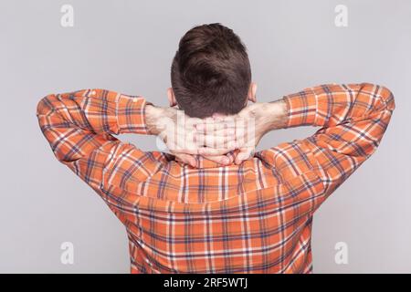 Portrait of dark haired man standing backwards to camera, raised his arms, holding neck, looking far, wearing orange checkered shirt. Indoor studio shot isolated on gray background. Stock Photo