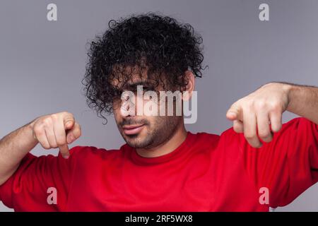 Portrait of funny attractive handsome young adult bearded man with curly hair, looking and pointing at camera, wearing red jumper. Indoor studio shot isolated on gray background. Stock Photo