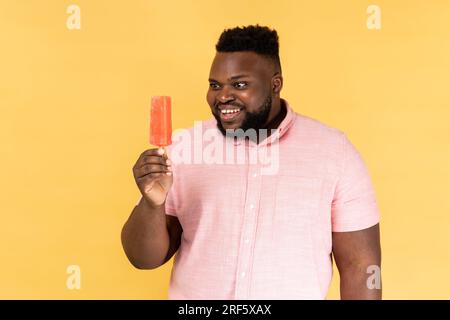Portrait of smiling man wearing pink shirt holding sweet ice cream wants to try delicious confectionery dessert in his hands. Indoor studio shot isolated on yellow background. Stock Photo