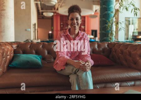 Smiling female freelancer sitting on sofa and looking at camera during working day in coworking Stock Photo