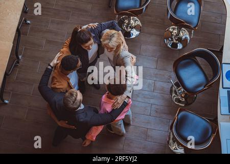 Top view of team of successful colleagues laughing and hugging while standing in circle in coworking Stock Photo