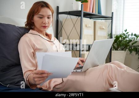Young transgender businesswoman sitting on bed in pajamas and working with documents Stock Photo